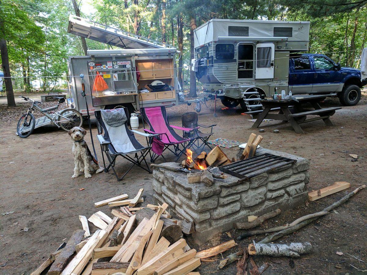 blue truck with with pop up camper next do a dog, chairs, bicicle, and fire pit